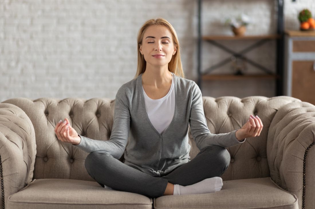unge gesunde schöne Frau üben Yoga zu Hause, sitzen in Lotus-Position auf dem Sofa, meditierend