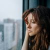Eine junge Frau mit braunen Haaren starrt nachdenklich und melancholisch durch ein vom Regen verschmiertes Fenster in einer Stadtwohnung, die depressiv aussieht