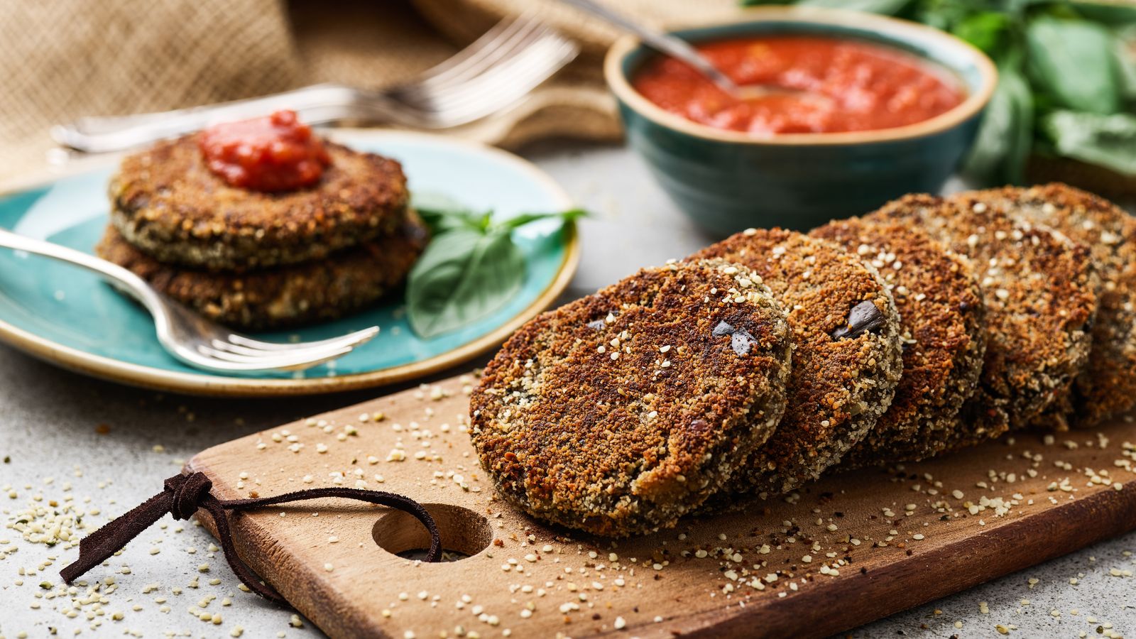 Burger mit Hanfsaat auf einem Holzbrett serviert. Im Hintergrund selbst gemachtes Tomatensugo in einer türkisen Schüssel
