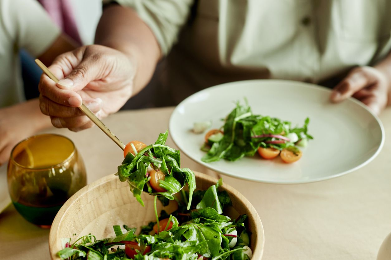 Hände einer schwarzen Frau, die Salat mit grünen Blättern auf den Teller am Tisch legt