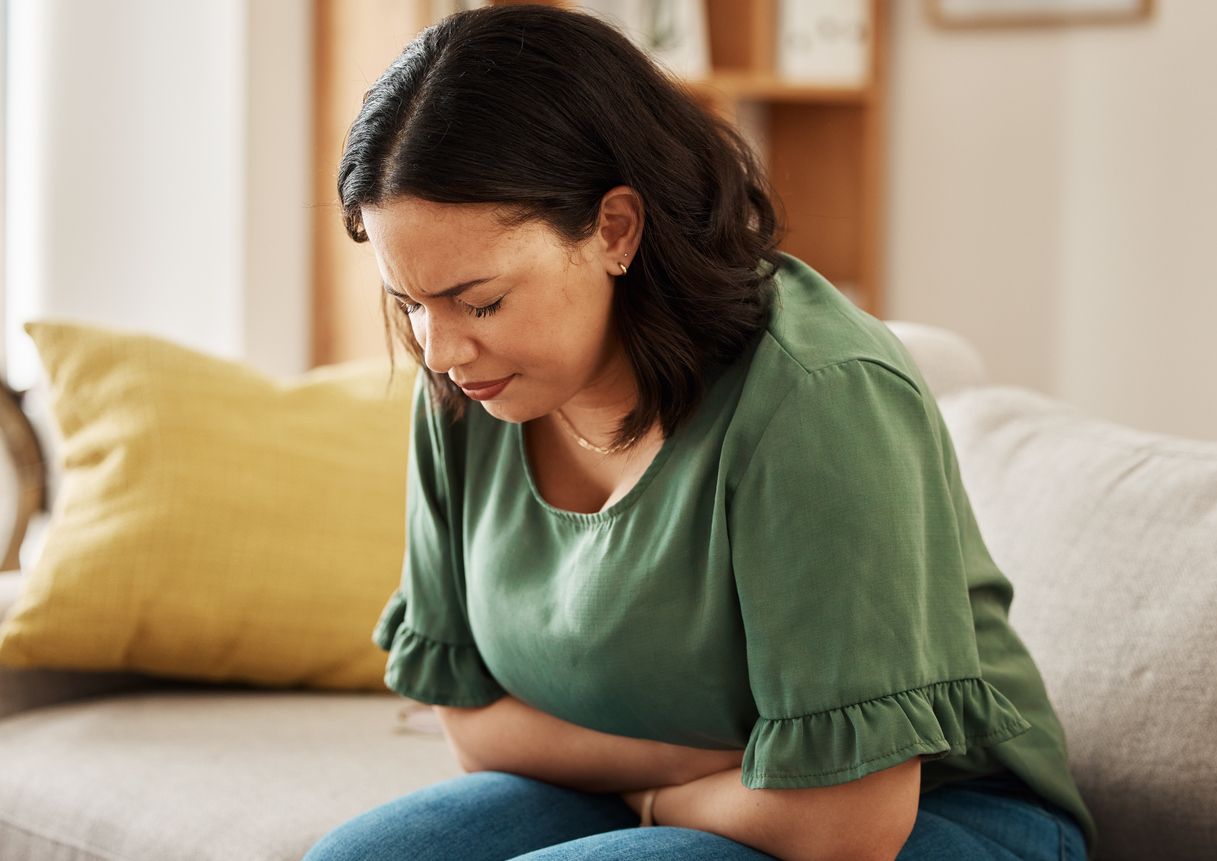Bauchschmerzen, Stress und Frau auf einem Sofa mit PMS, Blähungen oder Blähungen, Verstopfung oder Menstruationsübelkeit zu Hause